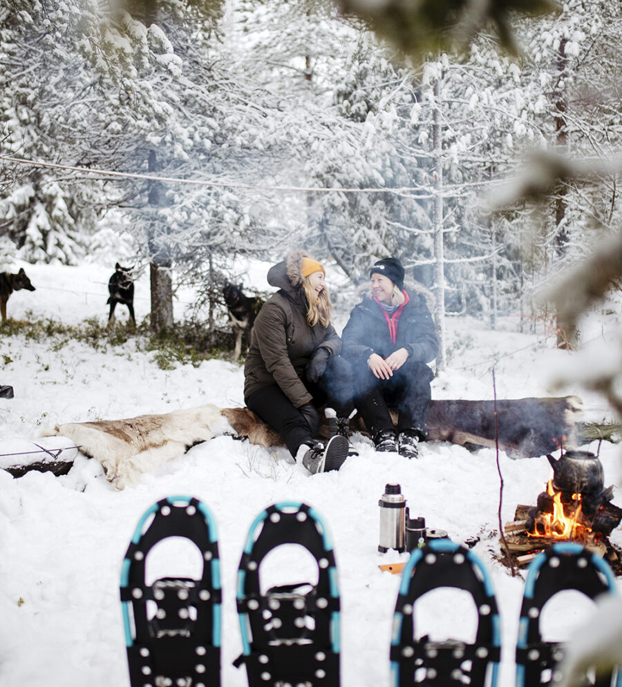 Two women talking and laughing beside a fire in Finnish Lapland after snowshoeing